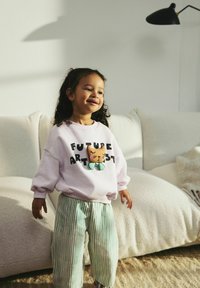 Jasnoróżowy sweatshirt z napisem "FUTURE ARTIST" i grafiką kota; zielone pasiaki z szerokimi nogawkami; dziecko stoi na pluszowym, jasnym dywanie.