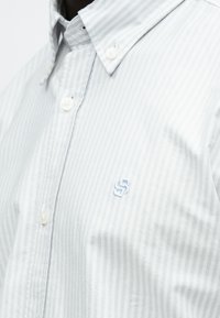Camisa de rayas gris claro con botones, hecha de algodón; cuenta con un cuello clásico y un pequeño logo bordado en azul en el pecho.
