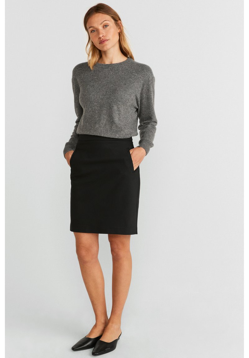 Pull en tricot gris à manches longues, associé à une jupe crayon noire avec poches latérales. Le modèle porte des mules noires à bout pointu.