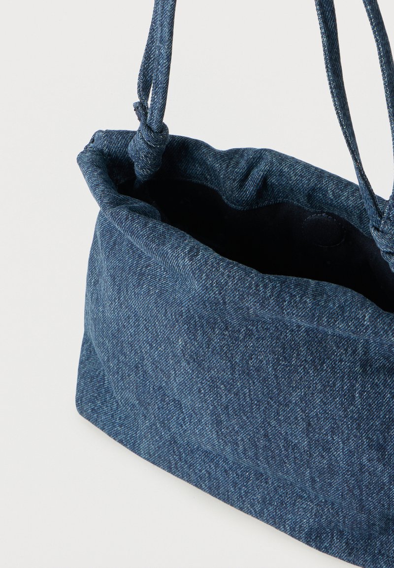 Borsa a tracolla in denim blu con coulisse, spallacci annodati e apertura superiore che mostra la fodera interna scura.