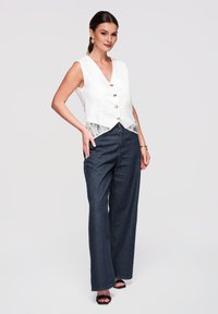 Femme portant un gilet blanc sans manches boutonné avec bordure en dentelle et un jean large noir, debout avec la main sur la hanche sur un fond uni.