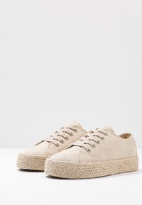 Beige kangaskengät, joissa on seitsemän nauhakanavaa, teksturoitu espadrille-tyylinen juuttialusta ja ommeltuja yksityiskohtia saumoissa.