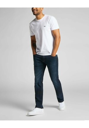 T-shirt en coton blanc avec un petit logo sur la poitrine, associé à un jean en denim foncé et des baskets blanches, offrant une silhouette décontractée et ample.