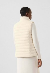 Gilet imbottito crema con colletto alto, caratterizzato da pannelli rettangolari trapuntati in orizzontale e una texture morbida, indossato sopra una maglietta a maniche lunghe coordinata.