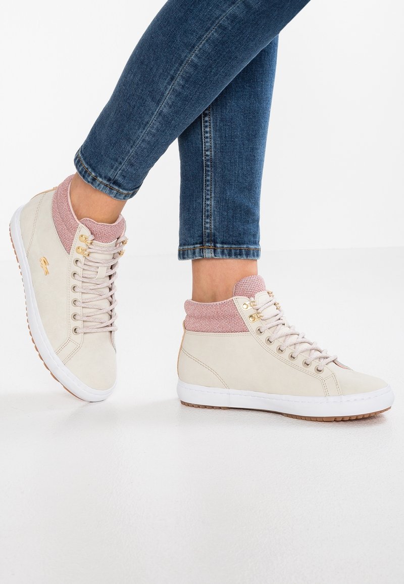 Zapatillas altas de gamuza beige con un cuello de punto rosa. Presentan ojales dorados y una suela de goma blanca con un agarre texturizado.