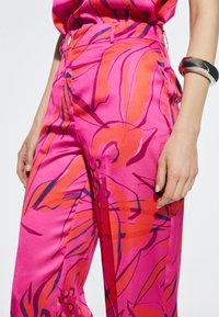 Roze satijnen broek met gedurfde bloemenpatronen in oranje en paars, met een hoge taille en zijzakken. Accenten zijn onder andere platte knopen aan de voorkant.