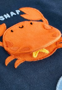 Orange broderad krabba med ett leende ansikte på en mörk marin bakgrund; har texturerade detaljer och gula accenter i sömmen.