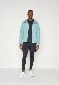 Lichtblauw waterdicht jack met een rits, zwart T-shirt met ronde hals, zwarte korte broek, zwarte legging en witte sportschoenen.