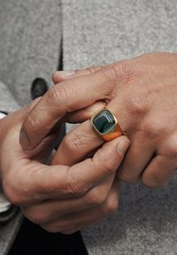 Gullring med en grønn rektangulær stein, teksturert ringstang, båret på hånden med synlige huddetaljer, foran en grå teksturert bakgrunn.