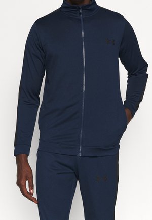 Homme portant une veste de sport bleu marine zippée intégralement et un pantalon assorti avec des logos Under Armour noirs sur la poitrine et la cuisse.