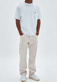 Hombre con camiseta blanca holgada, pantalones cargo beige con bolsillos laterales y zapatillas blancas con suelas beige en un entorno neutro.