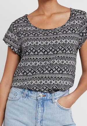 Person iført en sort og hvid kortærmet top med geometrisk mønster og lyseblå højtaljede jeans, hånden i lommen.