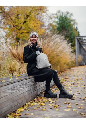 Žena v černém kabátě a masivních botách sedí na dřevěné lavici, drží světle šedý batoh, na zemi jsou podzimní listy, za ní vysoké trávy a stromy.