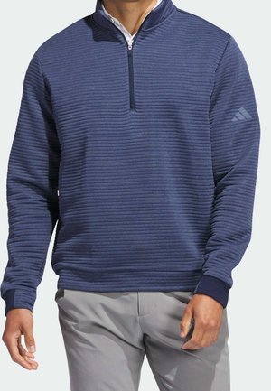 Homme portant un pull côtelé bleu marine à fermeture éclair quart de zip avec manches longues et un pantalon gris clair.