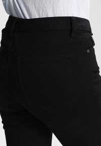 Vaqueros de mezclilla negros con una cintura alta, que presentan un bolsillo trasero con una costura diagonal, textura suave y un detalle de hardware mínimo.