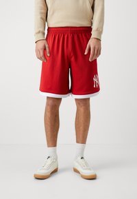 Shorts de sport rouges avec accents blancs, taille élastique et texture en maille ; présentent un logo blanc sur la jambe gauche. Portés avec des chaussettes blanches et des baskets.