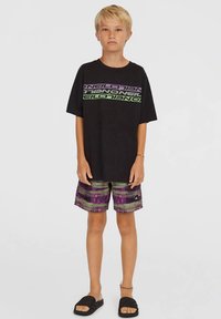 Černé bavlněné tričko s fialovým a zeleným logem, doplněné multikulturními šortkami se svislými pruhy a bočním logem, nošeno s černými sandály.