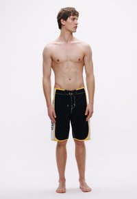 Černé boardshorty se žlutými akcenty a kontrastními bílými detaily. Mají elastický pas se stahovací šňůrkou a boční kapsy.