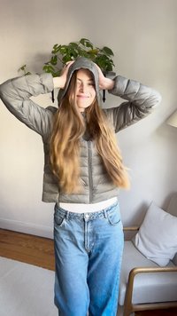 Gray puffer jacket with hood, featuring a zip front and quilted design, paired with light blue jeans and a white top. Indoor setting.