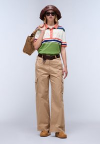 Veelkleurig gestreept poloshirt in groen, oranje en wit, gecombineerd met tan cargobroek met zijzakken en een bruine riem. Bruine emmerhoed en zonnebril.