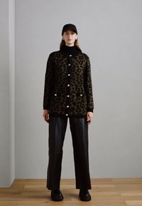 Gebreide cardigan met luipaardpatroon in olijfgroen en zwart, met grote knopen en een ronde kraag, gecombineerd met zwarte leren wijde broek en plateau schoenen.