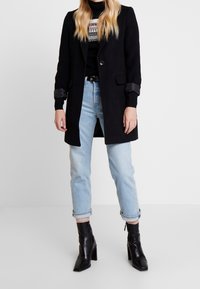 Svart ullblandad kappa med en enkel knapp, långa ärmar och manschettdetaljer, kombinerad med ljusblå uppvikta jeans och svarta klackiga ank stövlar.