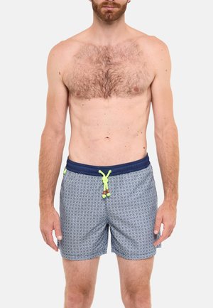 Homme torse nu avec poils sur la poitrine portant un short de bain bleu à motifs avec ceinture bleu marine et cordon jaune fluo sur fond blanc.