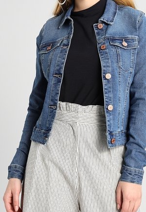 Femme portant une veste en jean bleue sur un haut noir, associée à un pantalon blanc taille haute avec de fines rayures verticales noires.