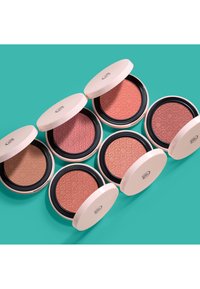 Cinq poudriers ronds avec couvercles beige et motifs embossés, présentant différentes nuances de poudre rose et pêche sur un fond bleu sarcelle.