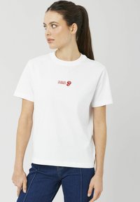 T-shirt blanc en coton avec col rond, manches courtes, et un petit logo rouge imprimé "The Rolling Stones" sur la poitrine.