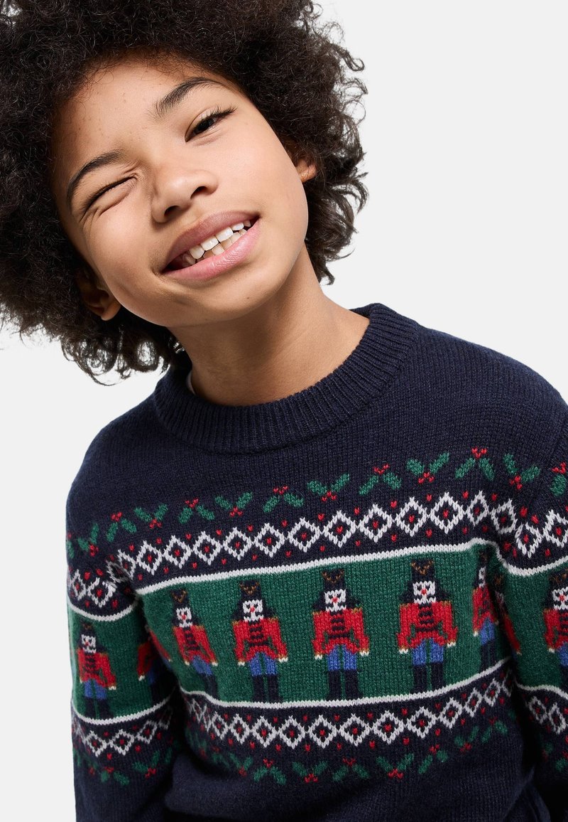 Pull bleu marine à col rond, orné d'un motif festif de casse-noisette en rouge et vert, agrémenté de motifs de houx et de losanges.