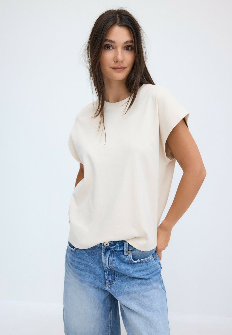Young woman with long dark hair wearing a loose white sleeveless top and blue jeans standing against a plain light background.
