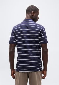 Άνδρας με πλάτη φορώντας μπλε σκούρο πόλο με λεπτές λευκές οριζόντιες ρίγες και μπεζ παντελόνι, μπροστά σε απλό φόντο.