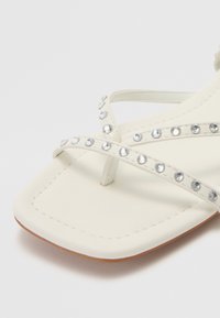 Des sandales plates blanches avec des lanières croisées doubles ornées de petits strass en argent, présentant un bout carré et une semelle plate.