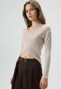 Lichtbeige geribbelde top met lange mouwen, een V-hals en een overslagontwerp, gecombineerd met donkerbruine, hoogheupse broek en een zwarte riem.