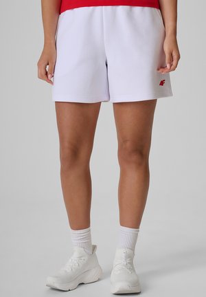Osoba ubrana w białe szorty sportowe z małym czerwonym logo, białe skarpetki typu crew, białe buty sportowe i czerwony top, stojąca na szarym tle.