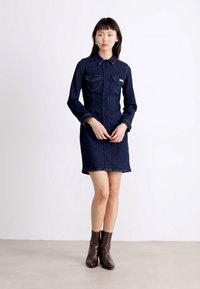 Jeanskjortekjole i mørkeblått, med krage, to brystlommer, knappestolpe foran og lange ermer. Modellen har på seg brune ankelstøvler.