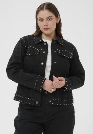 Vrouw die een zwart spijkerjack met studs draagt over een wit topje, staand tegen een effen lichte achtergrond, met handen samengevouwen ter hoogte van de taille.