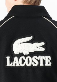 Jachetă neagră cu un logo proeminent brodat în alb, reprezentând un crocodil, și textul "LACOSTE" dedesubt, accentuată de un guler alb cu piping.