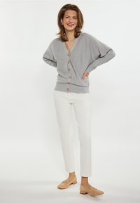Cardigan tricotat de culoare gri deschis, cu decolteu în V, nasturi din lemn și textură în relief, asortat cu pantaloni albi mulați și pantofi plate bej.