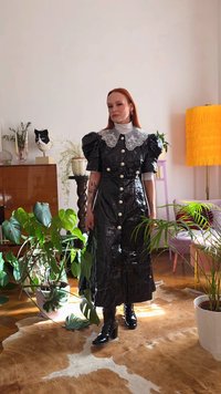 Vestido preto de plástico com mangas bufantes e uma gola de renda branca, com botões frontais. Usado com botas pretas de salto, rodeado de plantas.