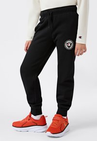 Zwarte fleece joggers met een elastische tailleband, zijzakken en een cirkelvormige logopatch. Gecombineerd met levendig rode sport schoenen.