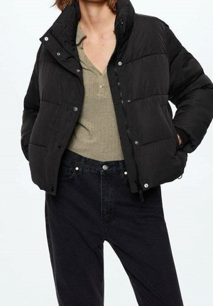 Personne portant une doudoune noire sur un t-shirt côtelé beige et un jean noir taille haute, les mains dans les poches de la veste.