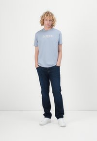 Vaaleansininen pyöreäkauluksinen t-paita, jossa on "GUESS"-logo paksuilla valkoisilla kirjaimilla, yhdistettynä tummiin farkkuihin ja valkoisiin lenkkareihin.