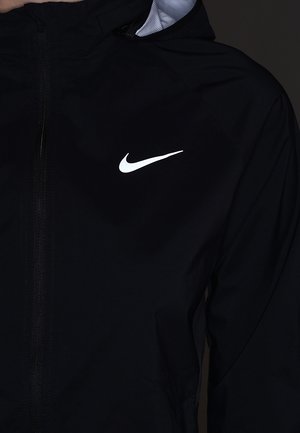 Schwarze Zip-Up-Jacke mit Kapuze und weißem Nike Swoosh-Logo auf der linken Brustseite vor dunklem Hintergrund.