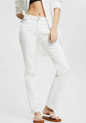 Kobieta w białych, prostych jeansach, białej koszuli zapinanej na guziki noszonej na czarnym krótkim topie oraz białych klapkach, stojąca na jednolitym tle.