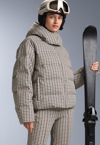 Padded ski jakna u hounds-tooth uzorku s visokim ovratnikom, uparena s odgovarajućim hlačama. Model drži crne skije. Nositi bijelu kacigu i naočale za skijanje.