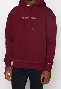 Felpa borgogna in cotone con tasca frontale. Presenta il logo "Tommy Jeans" in bianco e rosso, e dettagli nei polsini con il marchio.