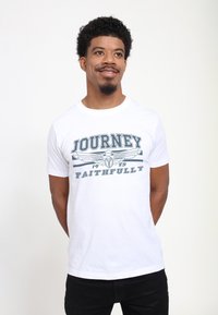 Usmívající se muž s krátkými kudrnatými vlasy, oblečený v bílém tričku s nápisem "Journey 1983 Faithfully" a křídleným emblémem, stojící s rukama za zády.