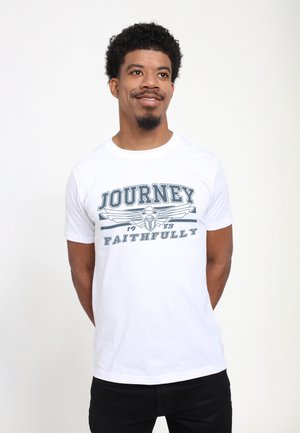 Smiling man with short curly hair wearing a white T-shirt with "Journey 1983 Faithfully" text and winged emblem, standing with hands behind back.